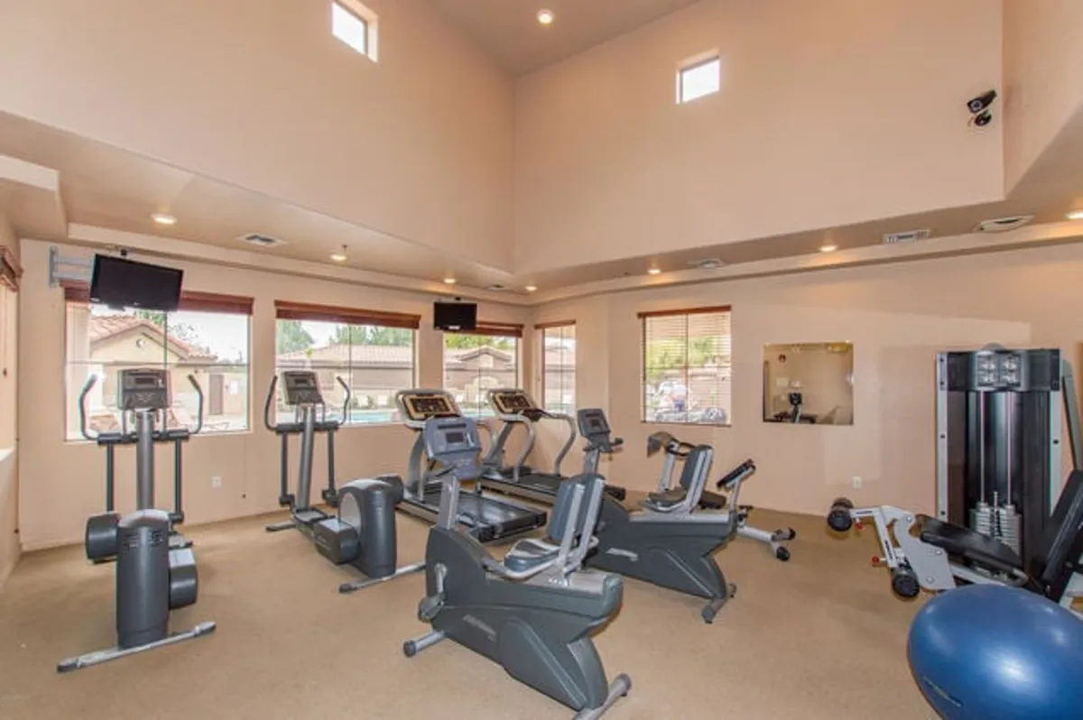 A bright, modern fitness room filled with treadmills, elliptical machines, and weight machines along large windows. The environment is active and motivating, reflecting community amenities for recreation and wellbeing.