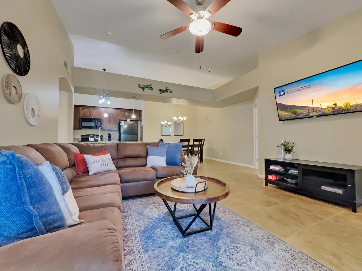 A spacious living room featuring a modern sectional sofa arranged around a round coffee table, a large TV displaying a scenic image, and stylish wall art. The room is bathed in natural light, suggesting proximity to large windows that might showcase mountain views.