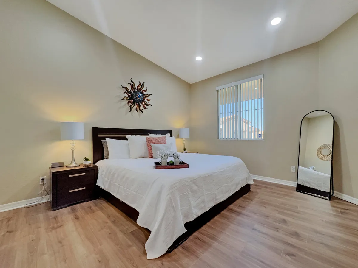A serene master bedroom with a bed featuring crisp white linens, decorative wall art above the headboard, matching nightstands with lamps on either side, and a large mirror reflecting soft natural light. The space feels airy and peaceful, designed for relaxation and restful sleep.