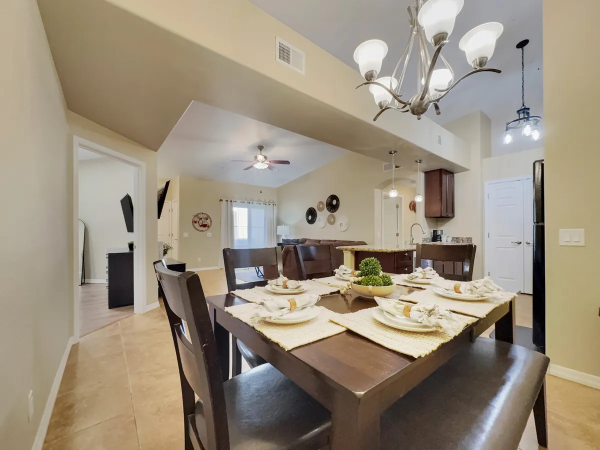 An open-plan dining space centered on a dining table fully set for a meal, accompanied by a contemporary chandelier and a view toward the adjacent kitchen. Modern appliances and finishes complete the inviting atmosphere.