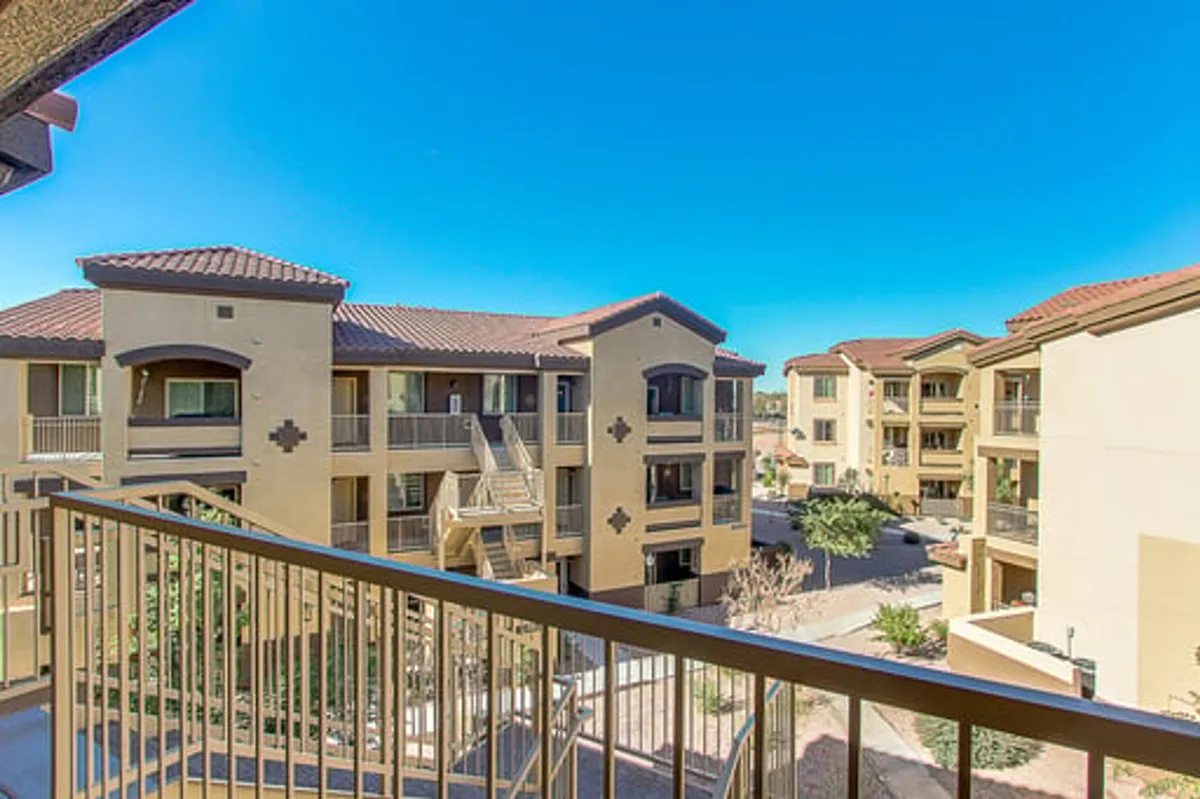 A private balcony with a railing overlooks surrounding buildings and a clear sky, with hints of mountain views in the distance. The scene evokes peaceful mornings or evenings spent outdoors, enjoying the scenery.