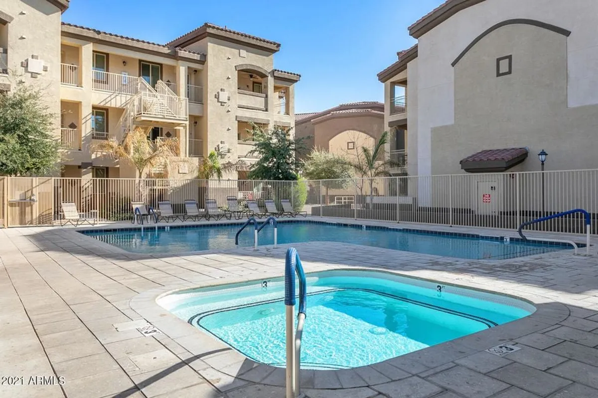 A sparkling swimming pool with an adjacent hot tub is surrounded by a row of inviting lounge chairs. The scene is set outdoors under blue skies, with lush landscaping at the edges of the pool area. The setting captures the core amenities of relaxation and recreation, perfect for unwinding alone or socializing with neighbors.