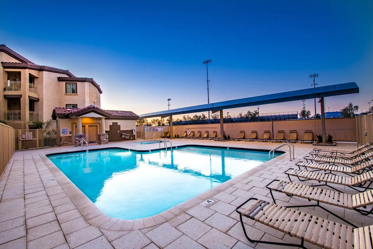 A tranquil swimming pool area featuring rows of poolside lounge chairs, a modern building facade in the background, and a shaded canopy over a seating area. The ambiance suggests a serene environment ideal for unwinding after a long day or gathering with friends.