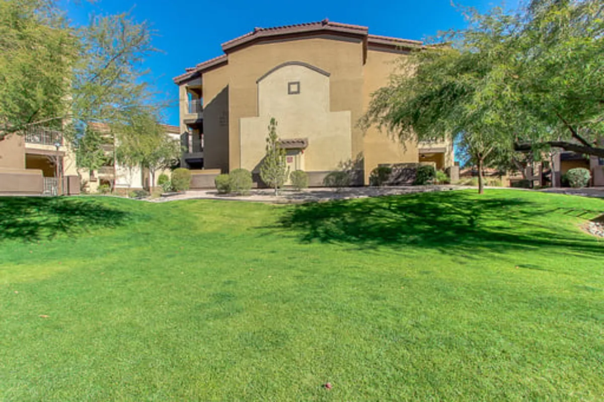 A peaceful exterior scene with a green lawn, well-kept landscaping, trees, and a view of the building's facade. The outdoor environment invites families to relax, gather for picnics, or enjoy the sunset from the balcony.