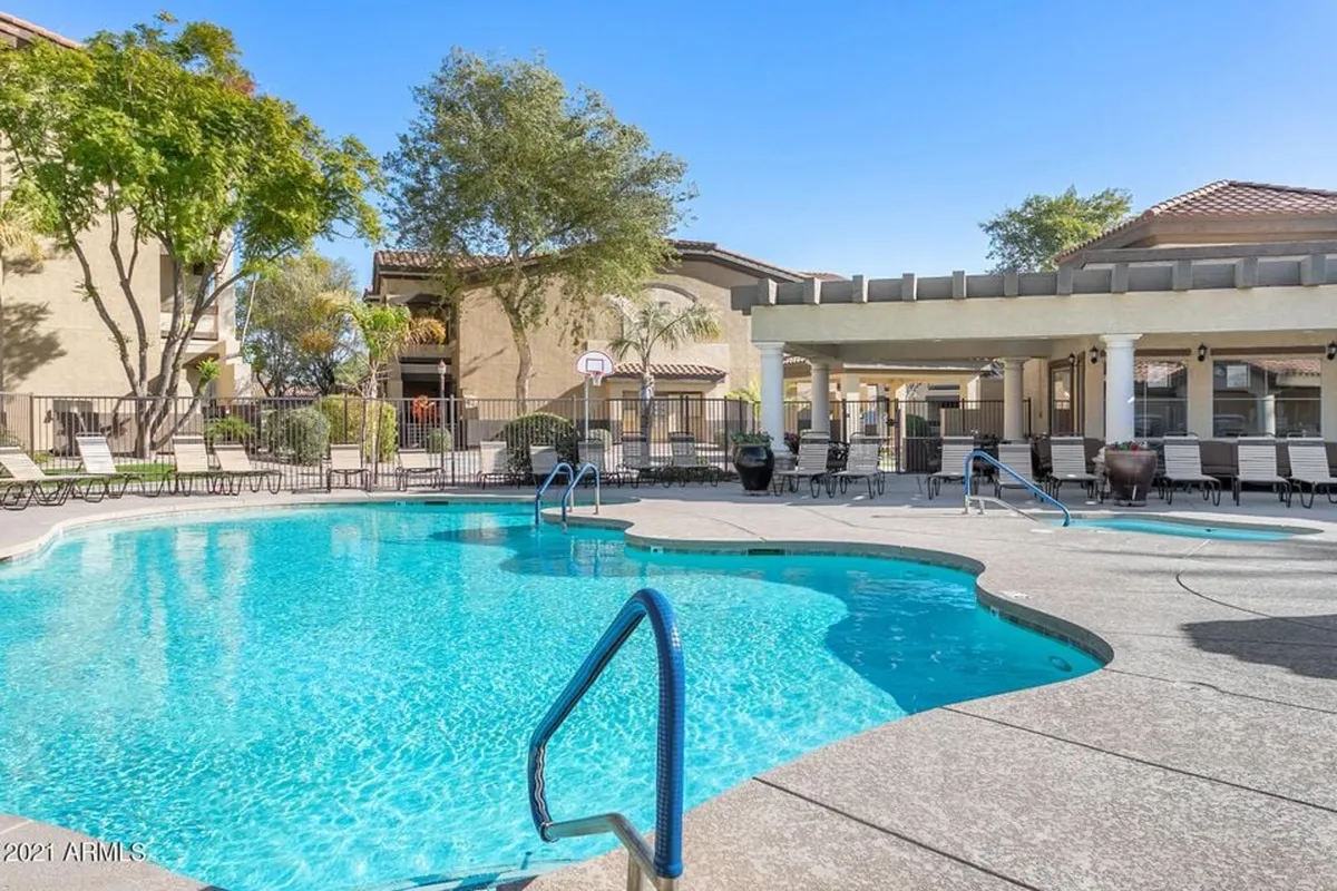 This image captures a large, resort-style swimming pool surrounded by lounge chairs, a covered patio area, and a basketball hoop, all set outdoors in a communal space. The pool area is inviting and signals leisure and activity opportunities for families.